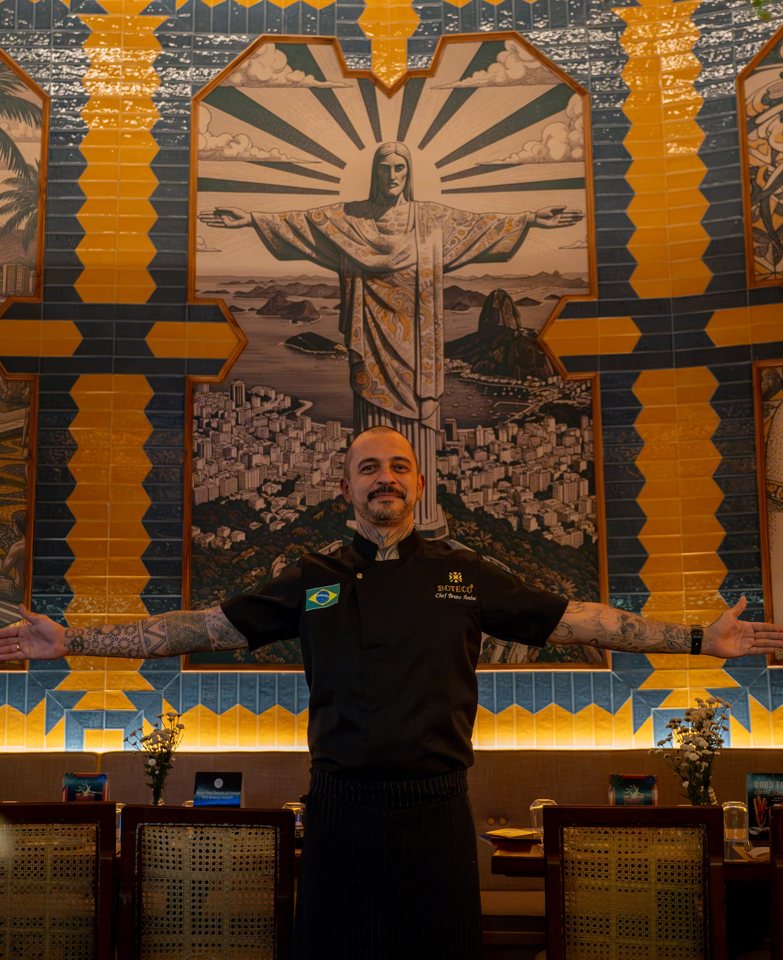 Chef Bruno Ambar with arms outstretched in front of Boteco's Brazilian mural